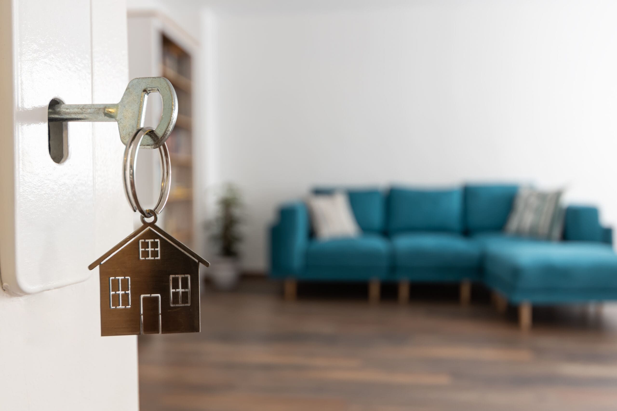 A key with a house-shaped keychain is inserted in a door lock, with a modern living room featuring a blue sofa and cushions blurred in the background—perfect for homes with recent kitchen installations or disabled adaptations.