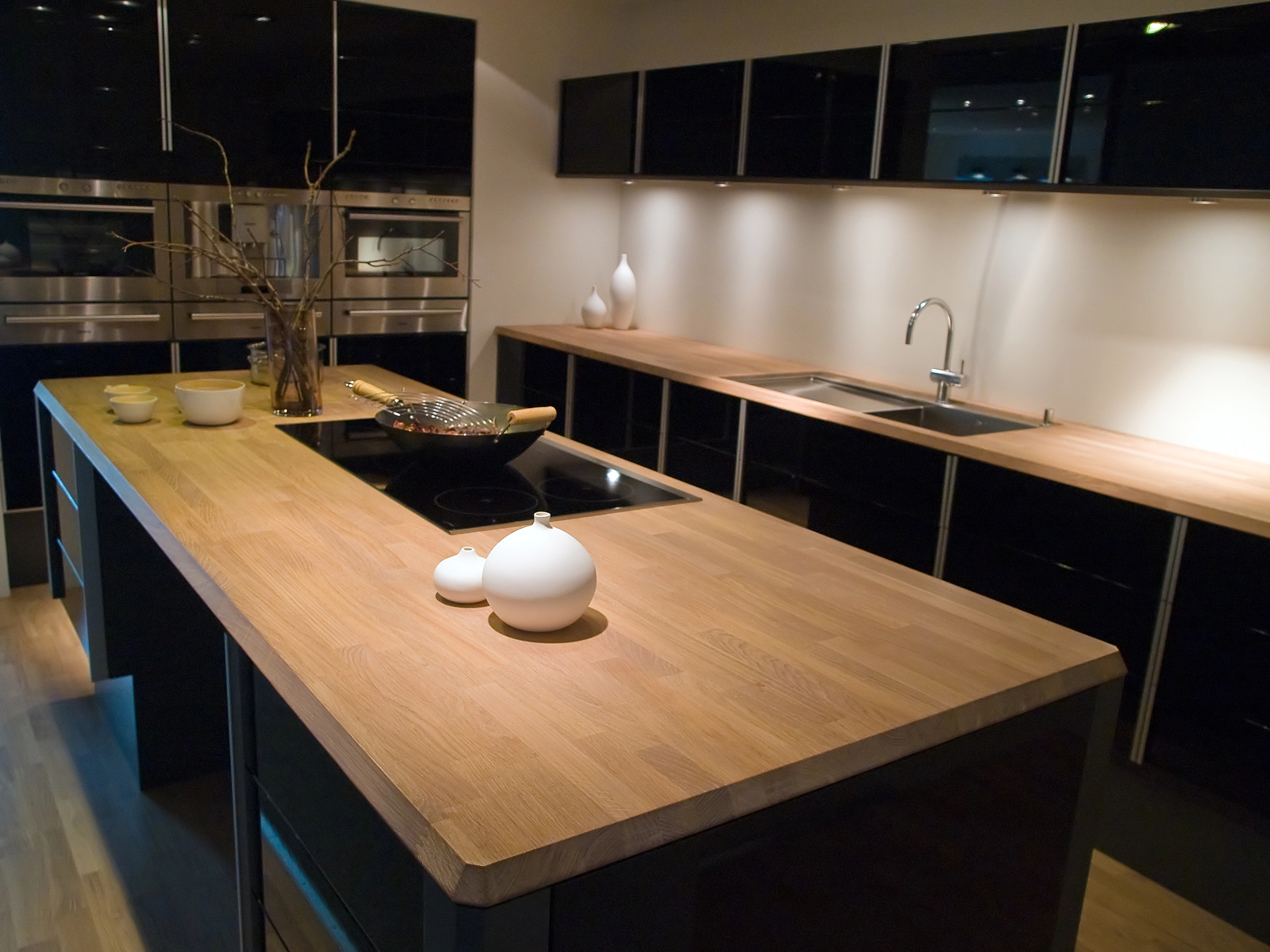 Modern kitchen with black cabinets, wooden countertops, built-in ovens, and an induction cooktop on a central island—ideal for stylish kitchen installations. Minimalist decor and soft, warm lighting create an inviting space.