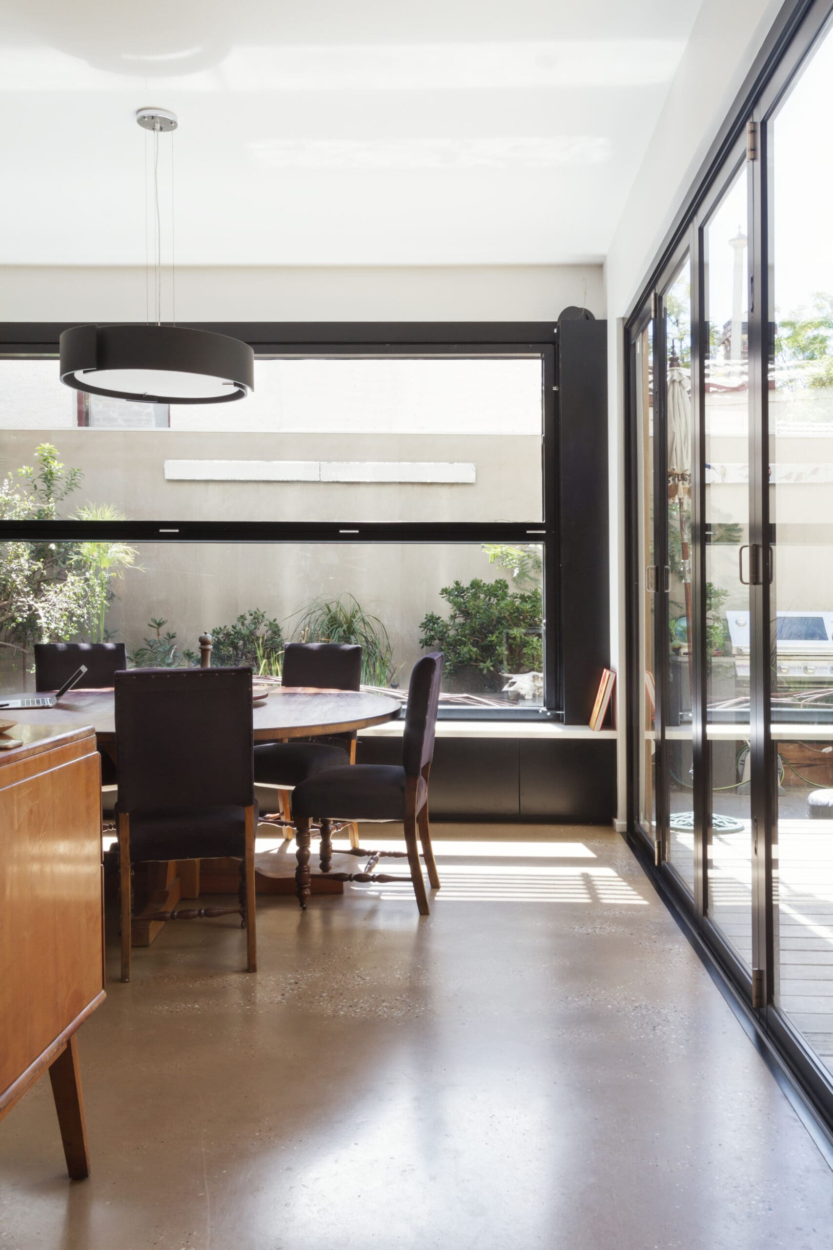 A modern dining area with a round table and dark chairs, large sliding glass doors, and abundant natural light. Perfect for house extensions, it features plants outside and a black circular pendant light above the table.