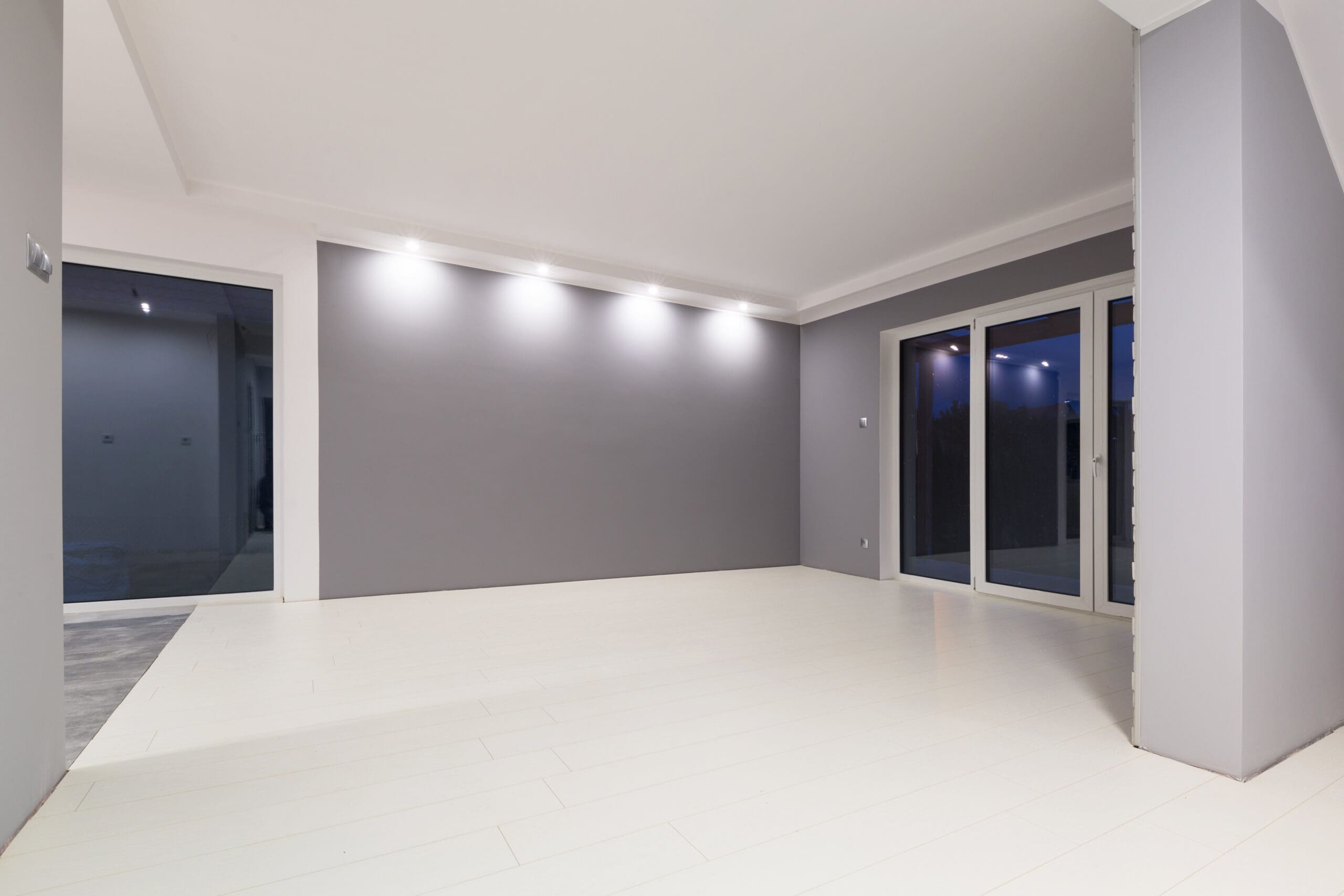 A modern, empty room with white flooring, gray walls, recessed ceiling lights, and large glass doors leading outside at dusk—showcasing the clean and minimalistic results of expert house extensions.