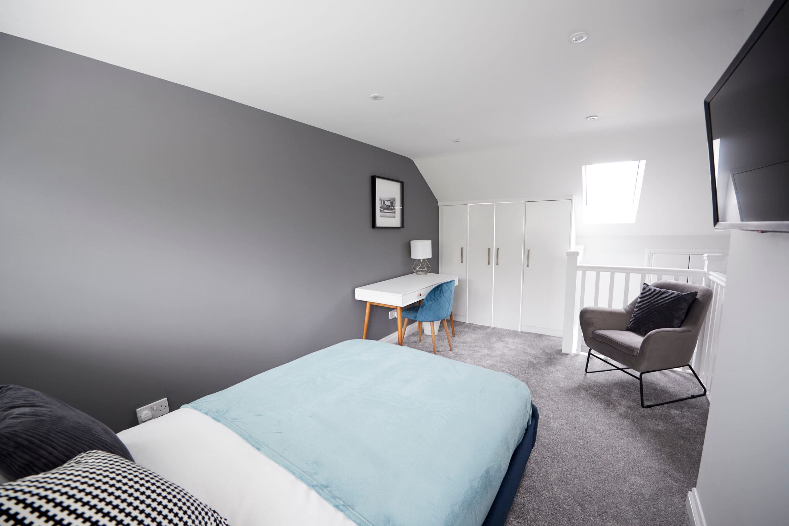 A modern bedroom with grey walls, a single bed with blue and white bedding, a white desk and chair, grey armchair, built-in white wardrobes, and a wall-mounted TV—perfect for homes after property refurbishments. Natural light enters from a skylight.