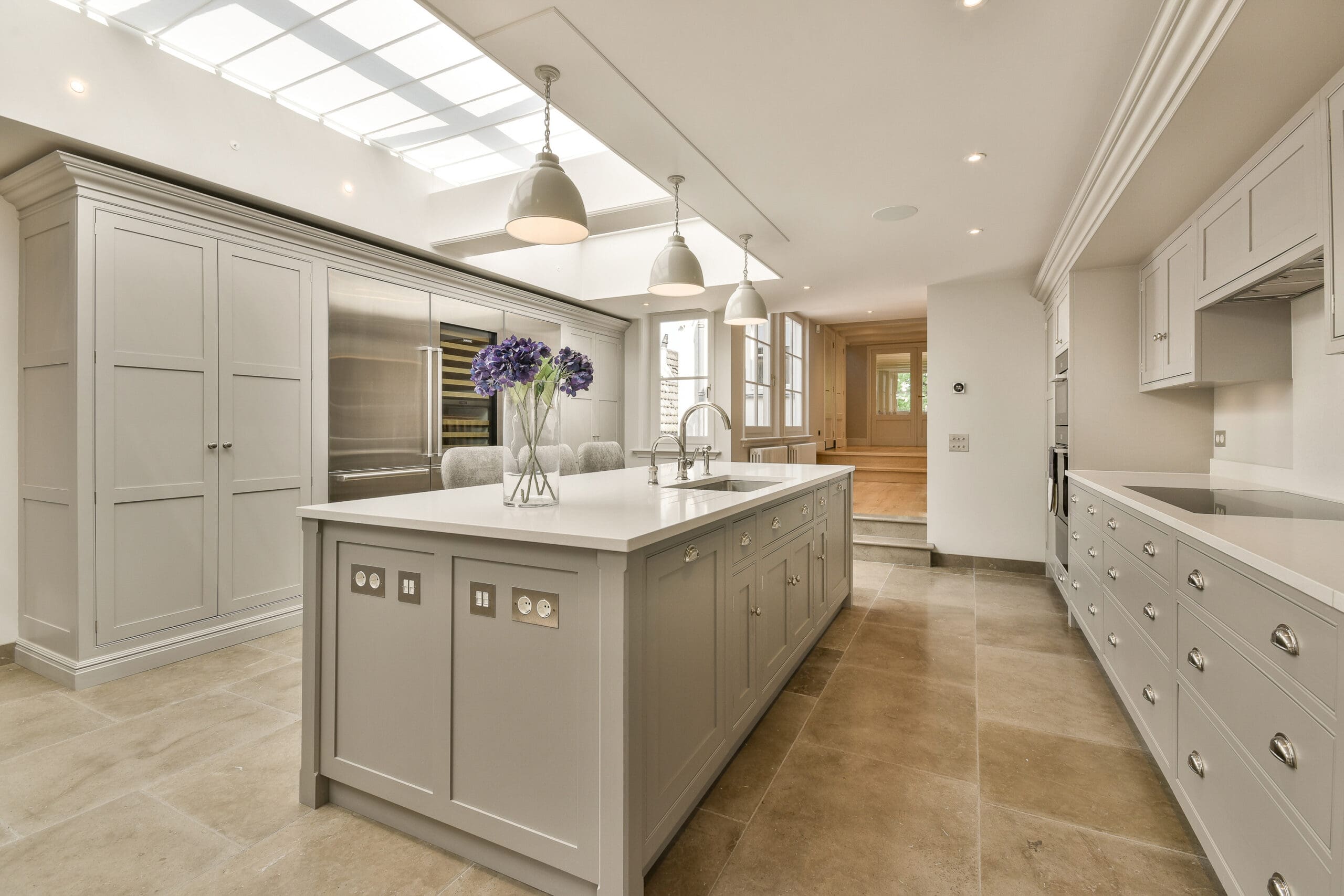 Modern kitchen with light grey cabinetry, large island with sink, bar stools, stainless steel appliances, pendant lights, skylight, and beige tiled floor. Expert in Kitchen Installations and House Extensions. A vase of purple flowers sits on the island.
