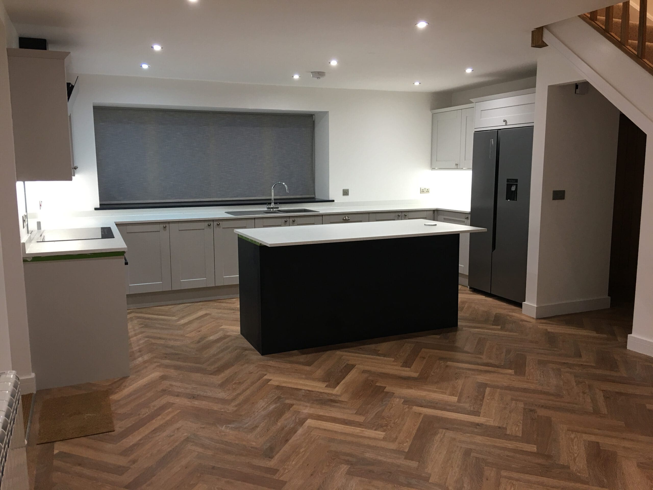 Modern kitchen with white cabinets, a dark central island, stainless steel double-door fridge, herringbone-patterned wood floor, and recessed lighting. Features include disabled adaptations and expert kitchen installations for added convenience.