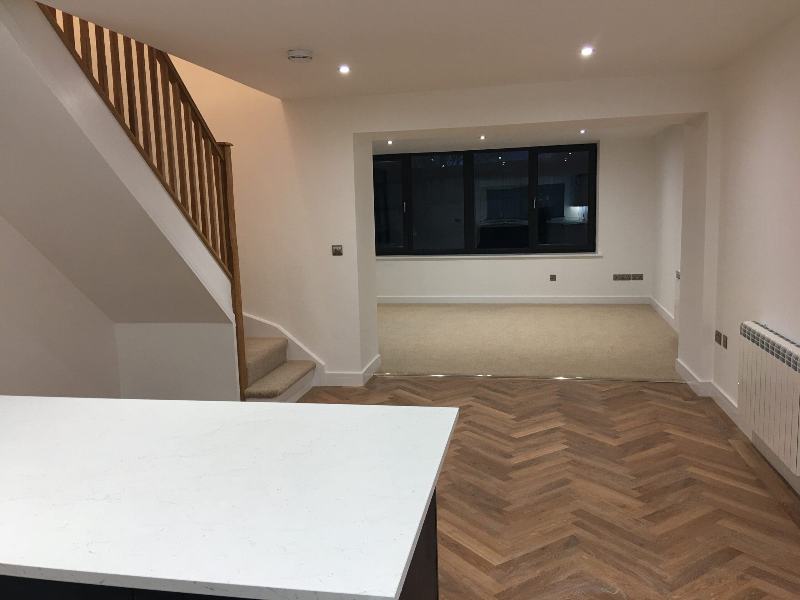 Modern, unfurnished open-plan living space with wood herringbone flooring, beige carpeted area, staircase with wooden bannister, large window, and recessed ceiling lights. Builder-quality kitchen installations visible in the foreground.