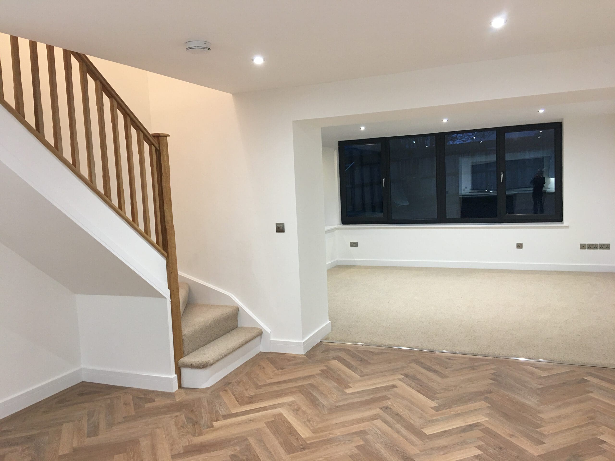 Modern, unfurnished living space with wood herringbone flooring, a carpeted area, a staircase with wooden railing, and large dark-framed windows. Perfect for those seeking quality builder work or planning kitchen installations.