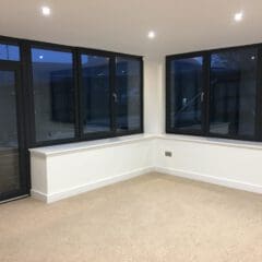 A bright, empty room with beige carpet, white walls, and black-framed windows showcases the quality of a skilled builder—perfect for house extensions or incorporating disabled adaptations. Recessed ceiling lights are on, and it appears to be evening outside.