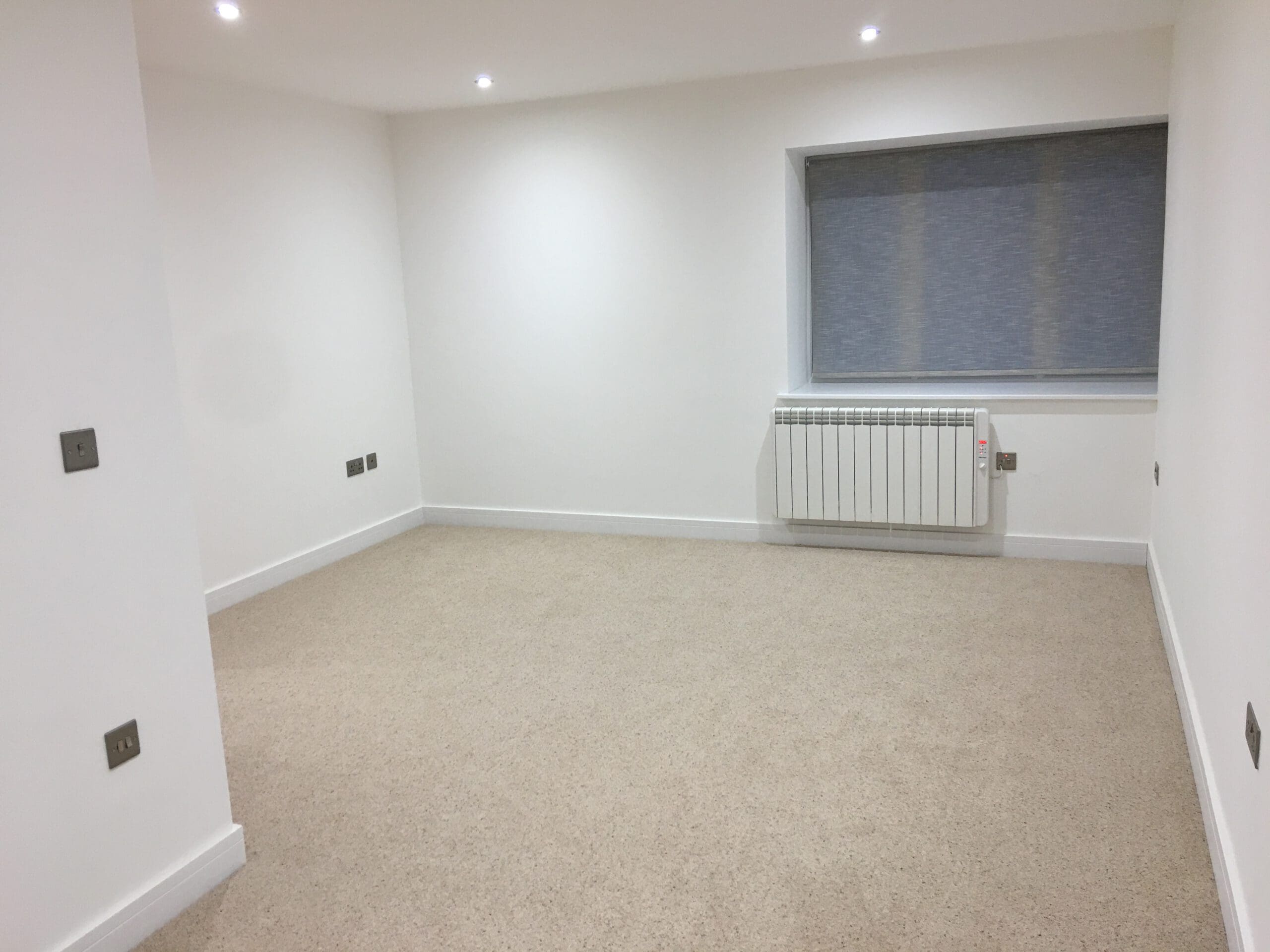 A clean, empty room with white walls, beige carpet, recessed ceiling lights, a radiator under a large window with a closed blue blind, and several electrical outlets—ideal for Property Refurbishments or future Kitchen Installations.