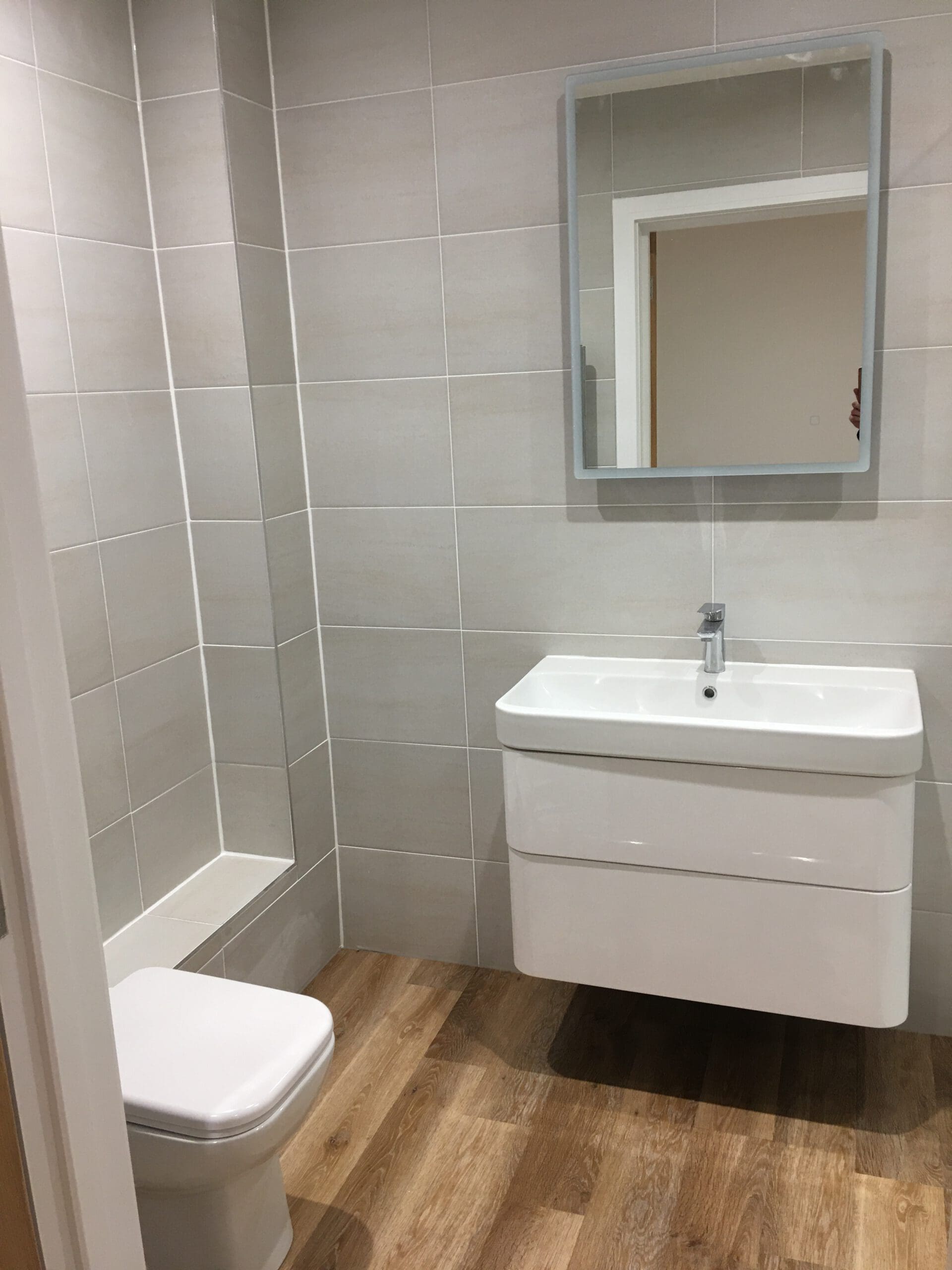 Modern bathroom with light beige tiles, wooden floor, a white toilet, built-in tiled bench, and a floating white sink with rectangular mirror above—ideal for disabled adaptations or property refurbishments by an experienced builder.