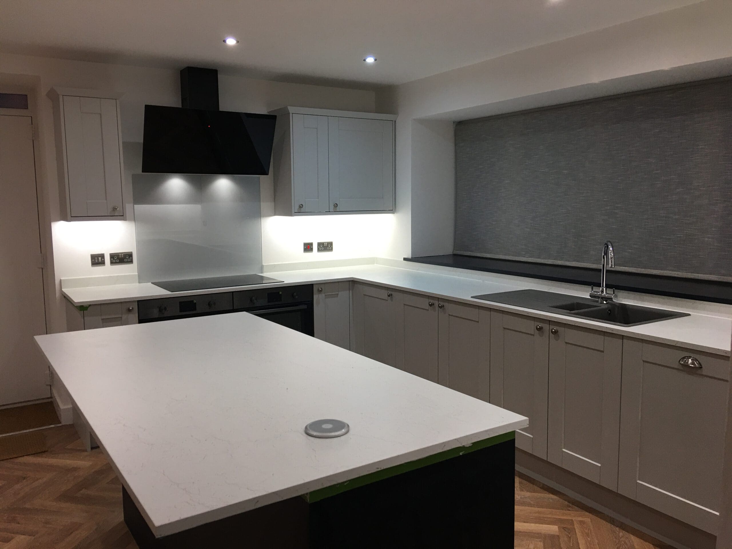 Modern kitchen with white cabinets and countertops, an island, built-in oven, black stove hood, stainless steel sink, and a large window with a grey blind—perfectly finished by an expert builder specializing in property refurbishments. Ceiling spotlights illuminate the space.