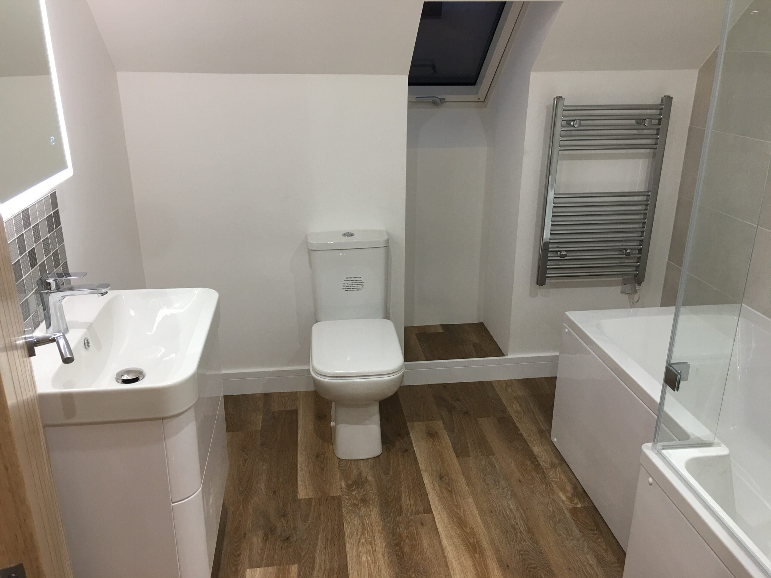 Modern bathroom with wooden flooring, white sink and cabinet on the left, toilet in the center, bathtub with glass screen on the right, wall-mounted towel radiator, skylight window above—ideal for homes needing Disabled Adaptations or House Extensions.