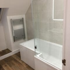 Modern bathroom with a white bathtub, glass shower screen, tiled wall, heated towel rail, and wood-style flooring under a sloped ceiling. Perfect for property refurbishments or as part of larger house extensions. Lighted mirror above the tub.