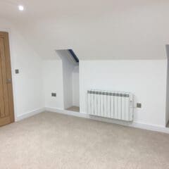 A bright, empty room with beige carpet, white walls, a wooden door, two small skylight windows, a white electric radiator, and sloped ceilings—perfect for house extensions or customized disabled adaptations by an experienced builder.
