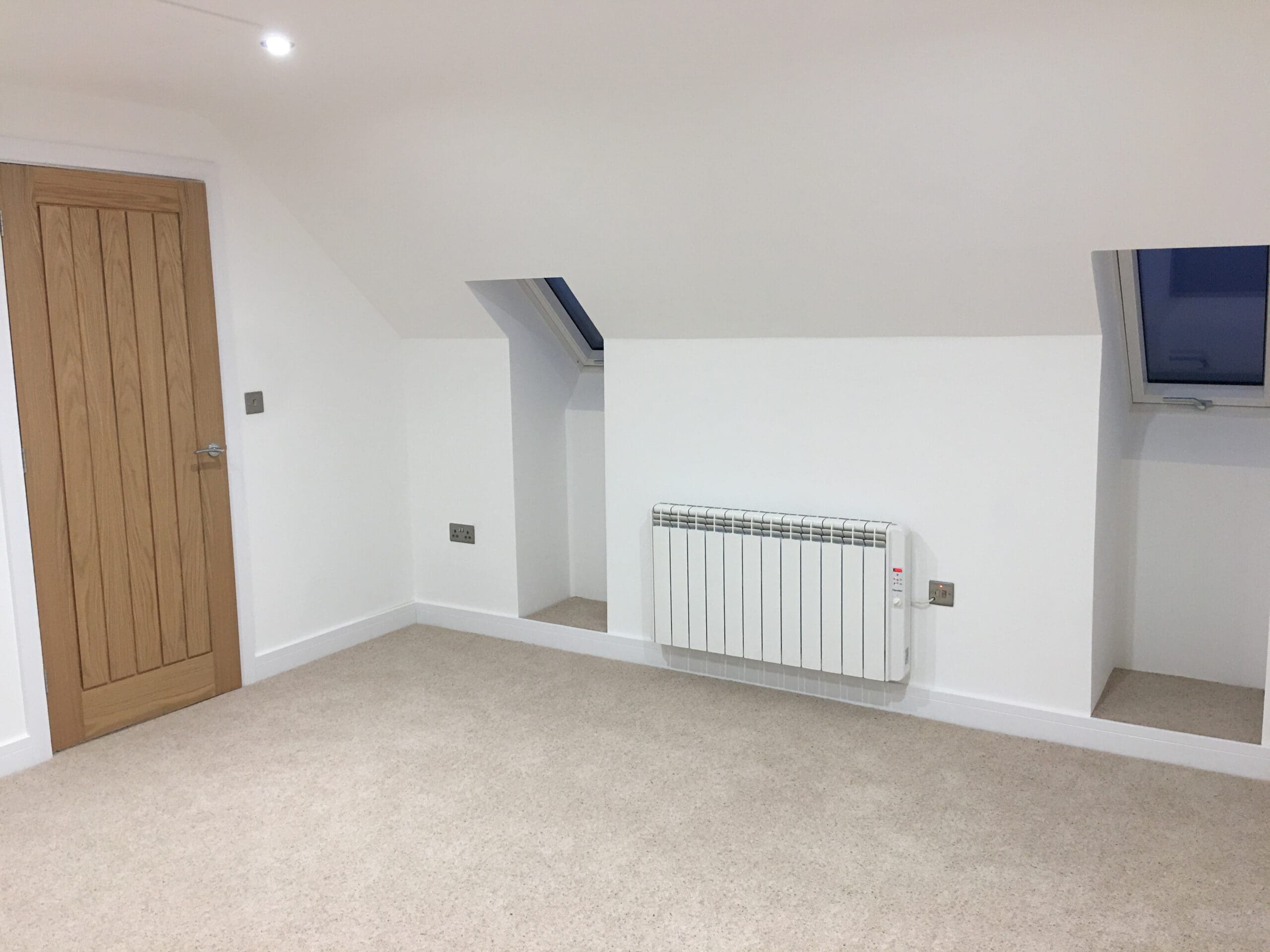 A bright, empty room with beige carpet, white walls, a wooden door, two small skylight windows, a white electric radiator, and sloped ceilings—perfect for house extensions or customized disabled adaptations by an experienced builder.