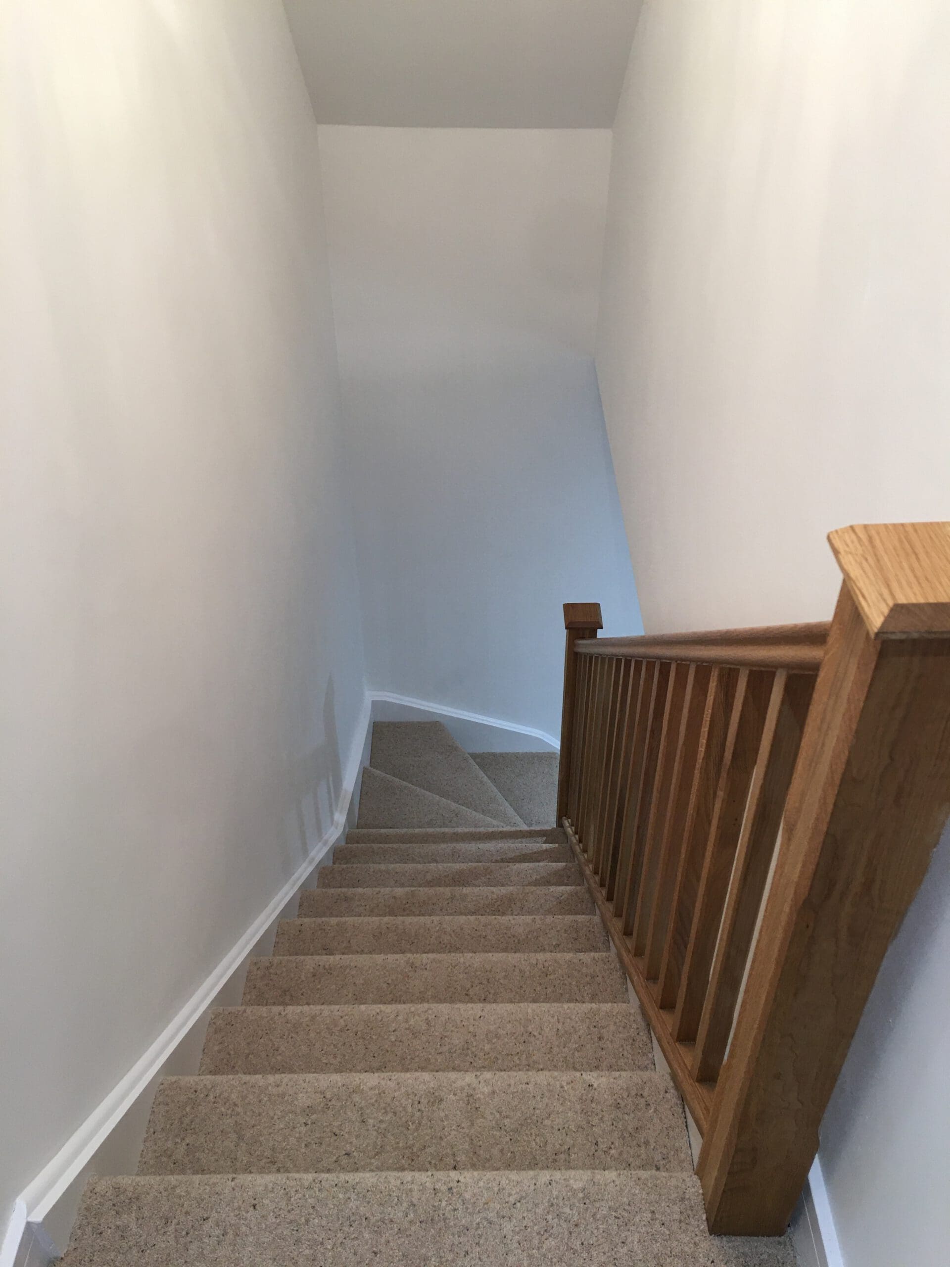 A carpeted staircase with a wooden handrail on the right leads down to a landing, surrounded by plain white walls—a classic feature often updated during property refurbishments. A small angled section sits at the bottom.