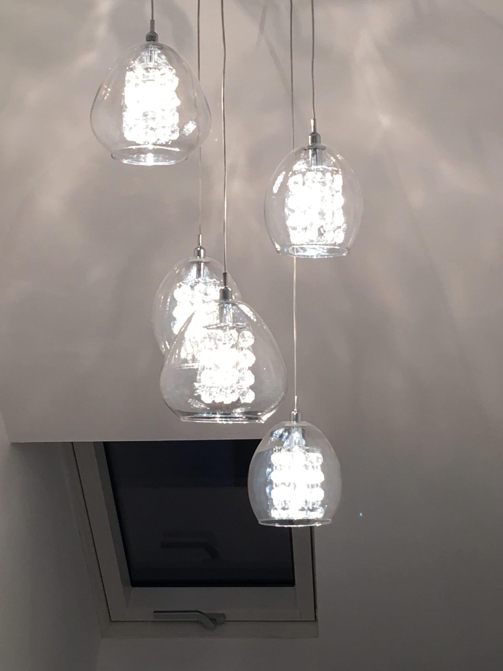 Five modern pendant lights with clear glass shades hang from the ceiling, glowing brightly. A rectangular window is partially visible below the lights—an elegant touch for kitchen installations or property refurbishments in contemporary homes.
