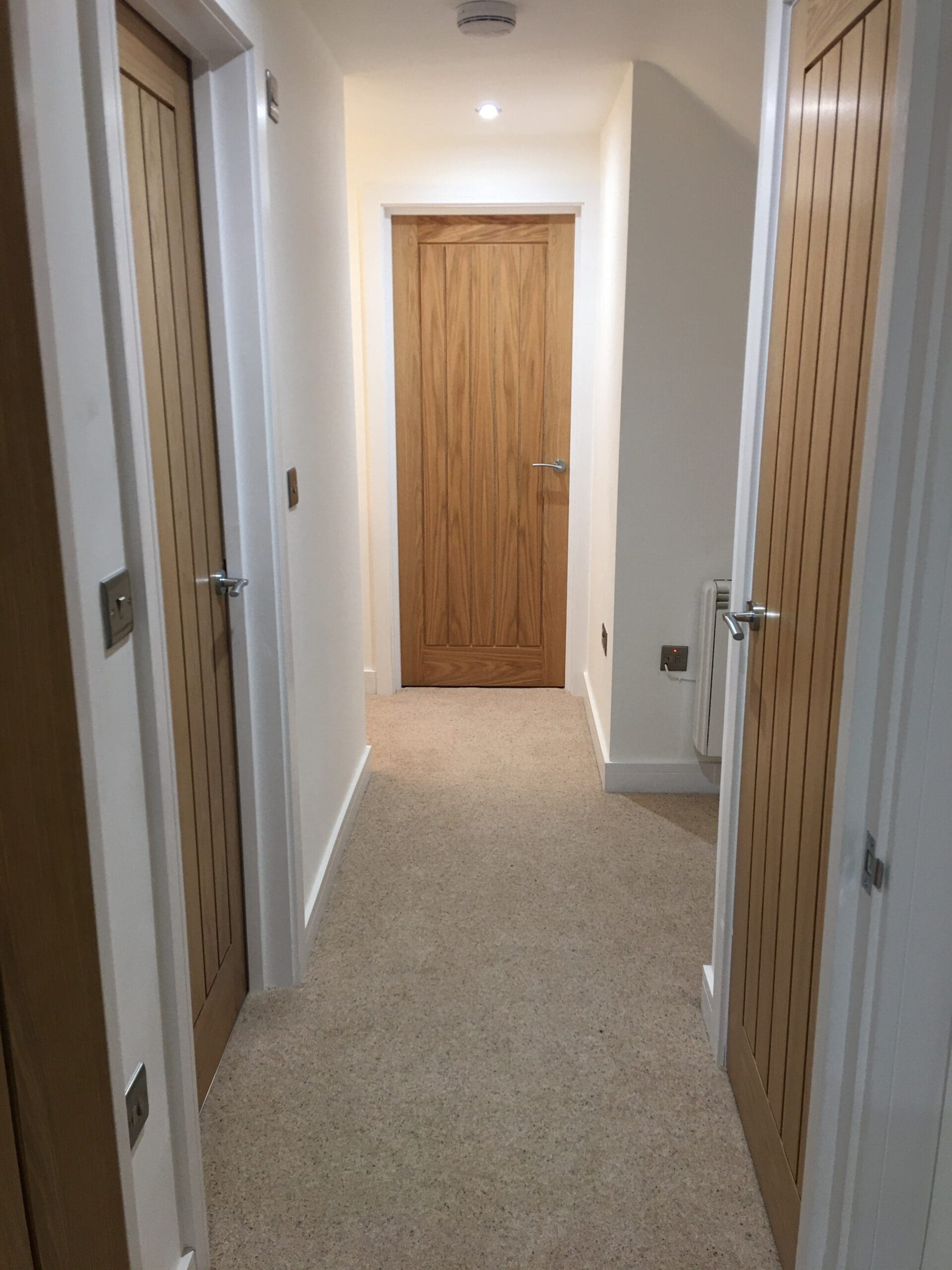 A hallway with beige carpet and white walls, designed by an expert builder, featuring three closed wooden doors—two on the sides and one at the end of the hallway.