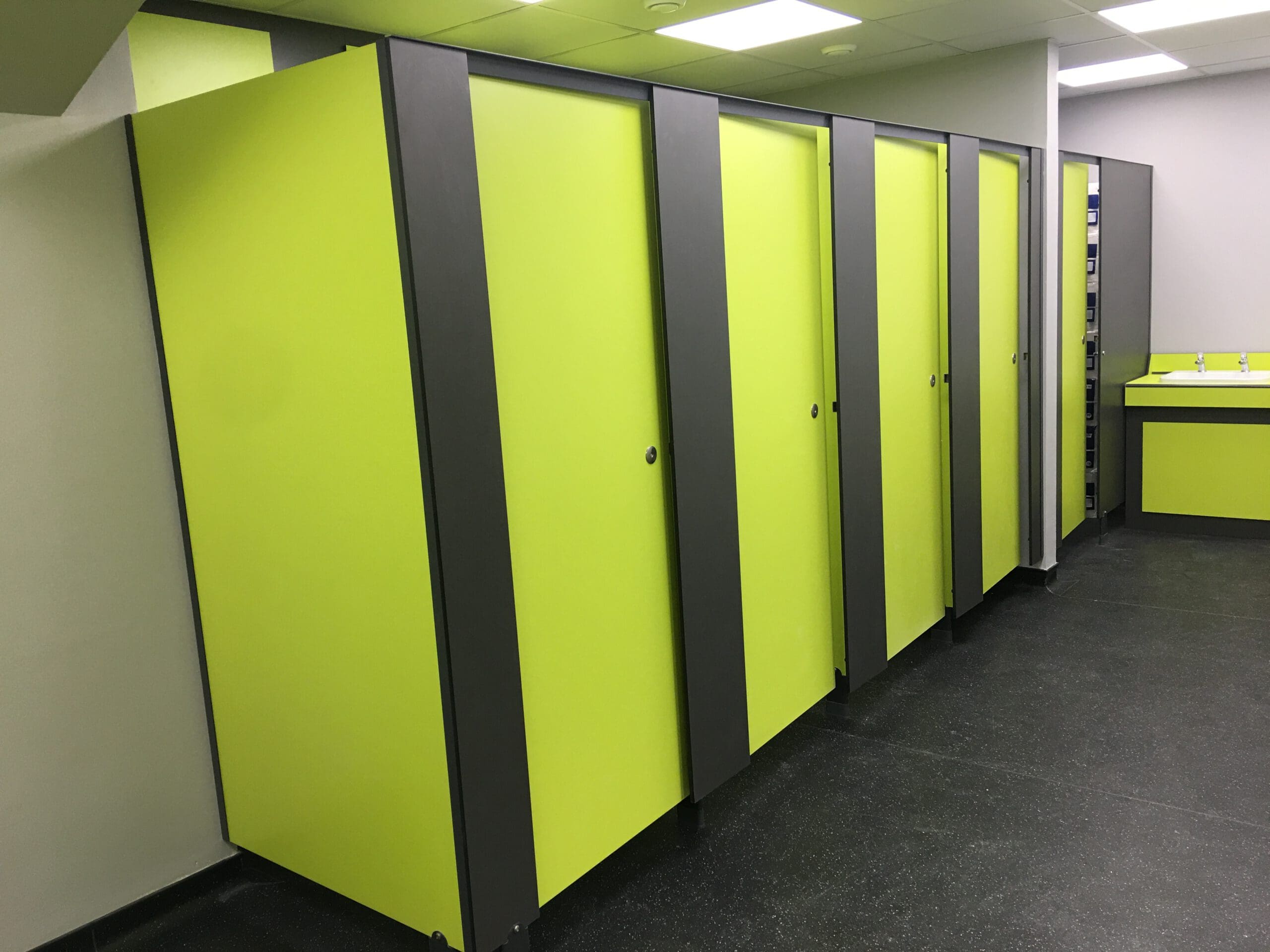 A row of green and gray restroom stalls with closed doors is shown, designed for Disabled Adaptations. To the right, a matching sink area with mirrors is visible. The floor is dark, and the walls and ceiling are light-colored.