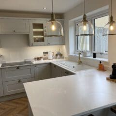 Modern kitchen with white countertops, grey cabinets, and three glass pendant lights. A window above the sink lets in natural light. Expert builder kitchen installations include utensils, a coffee machine, and a soap dispenser on the counters.