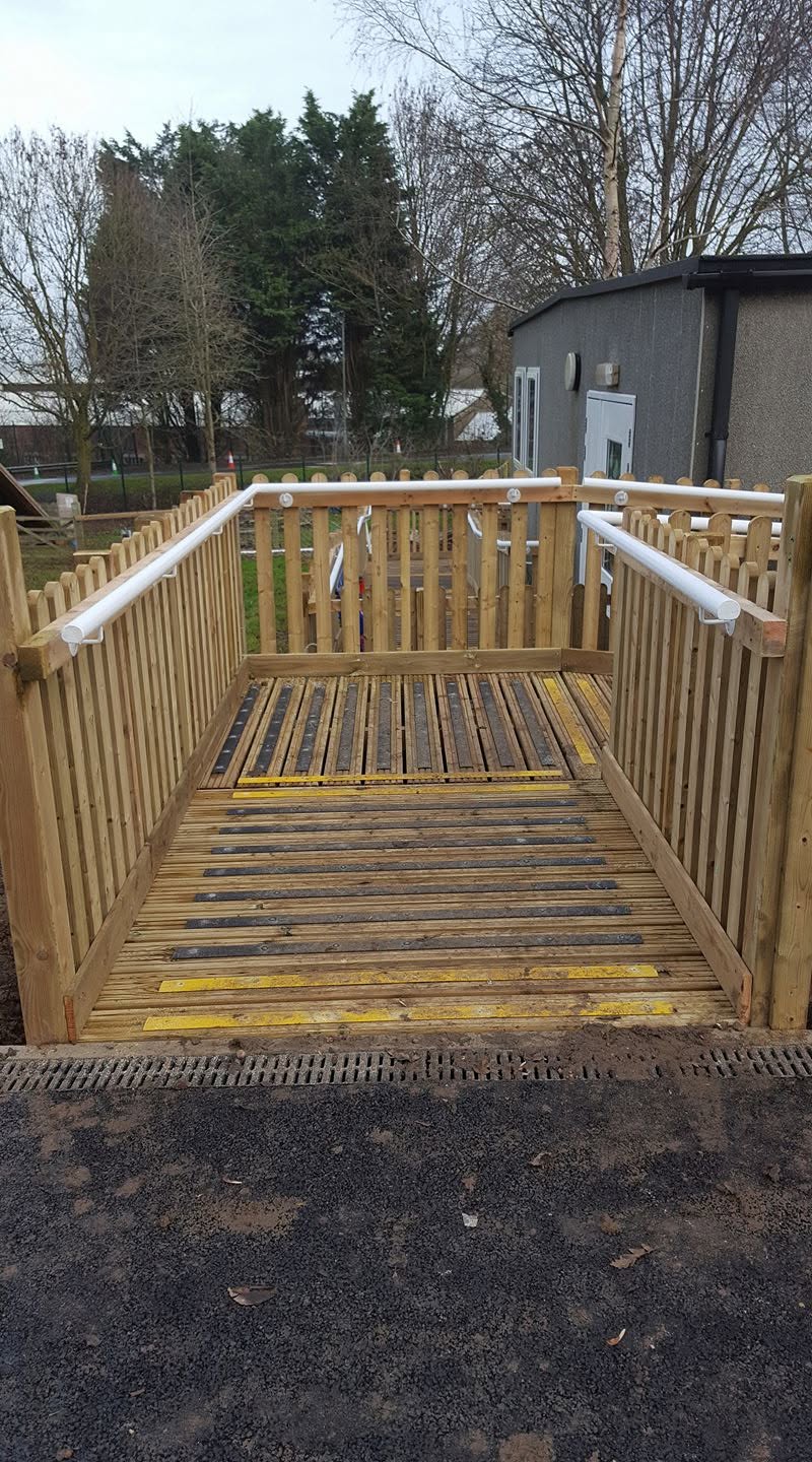 A wooden accessibility ramp with railings and white hand grips leads up to a building. The ramp has yellow safety markings and trees are visible in the background.