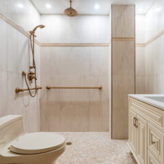 Accessible bathroom with a walk-in shower, handheld showerhead, grab bars, a toilet, and a white vanity with a sink, all featuring gold fixtures. The floor has small beige tiles and the walls are tiled.